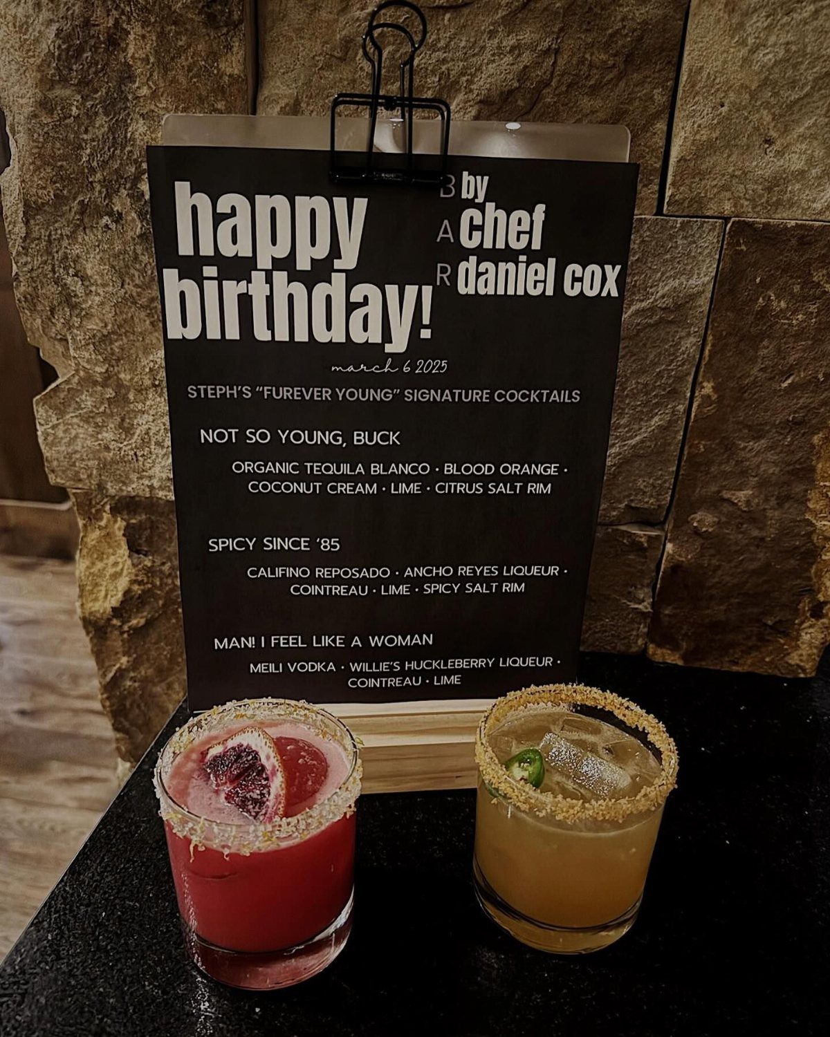 Two colorful cocktails with textured rims sit before a printed signature cocktail menu.