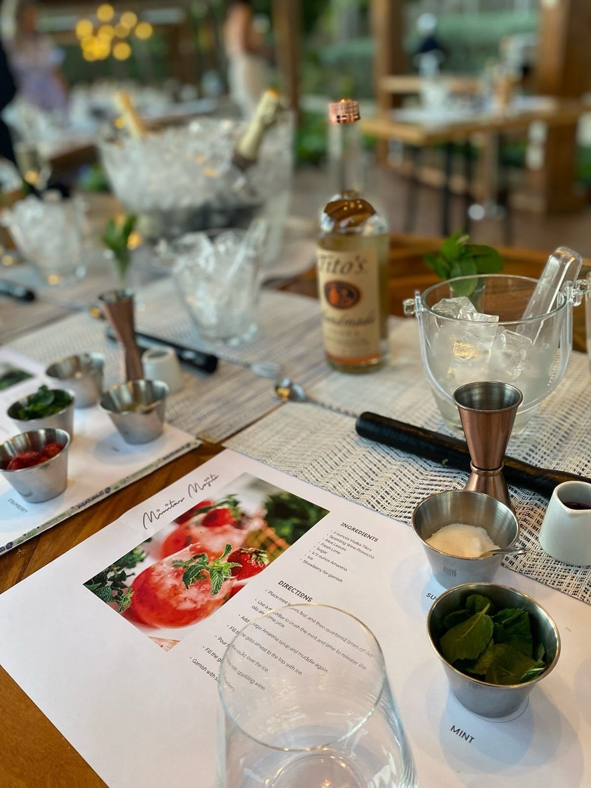 Cocktail making supplies including vodka, jiggers, ingredients in small metal bowls, and a recipe card on a table.