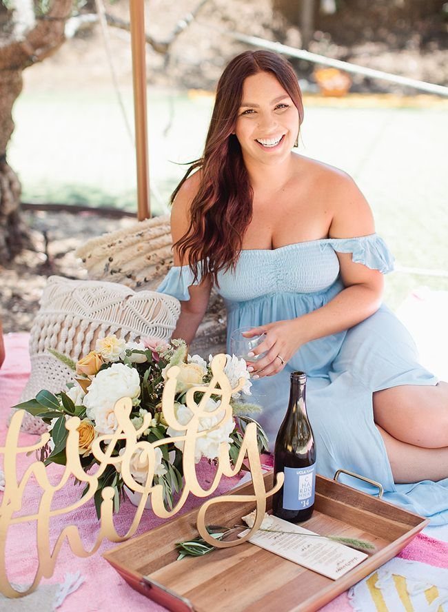 Smiling woman in a blue dress sitting on a pink picnic blanket with flowers and wine.