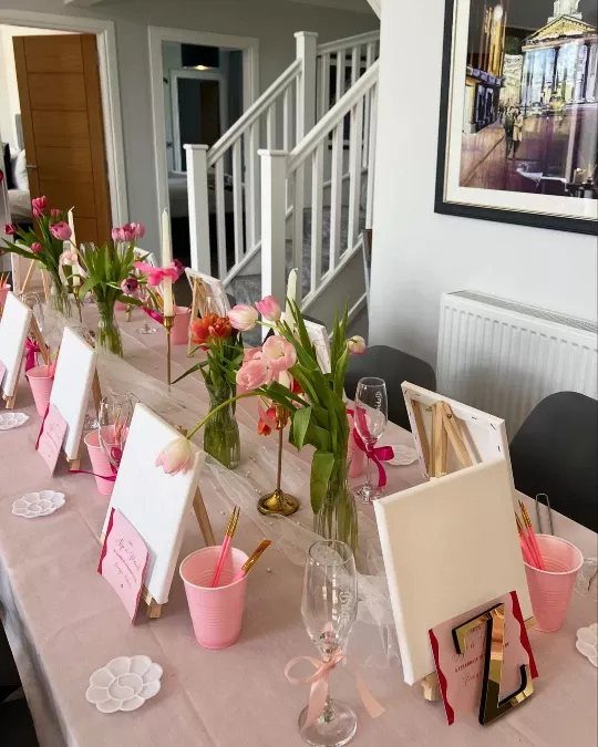 A long table set for a paint and sip party with canvases, pink cups, and tulips.