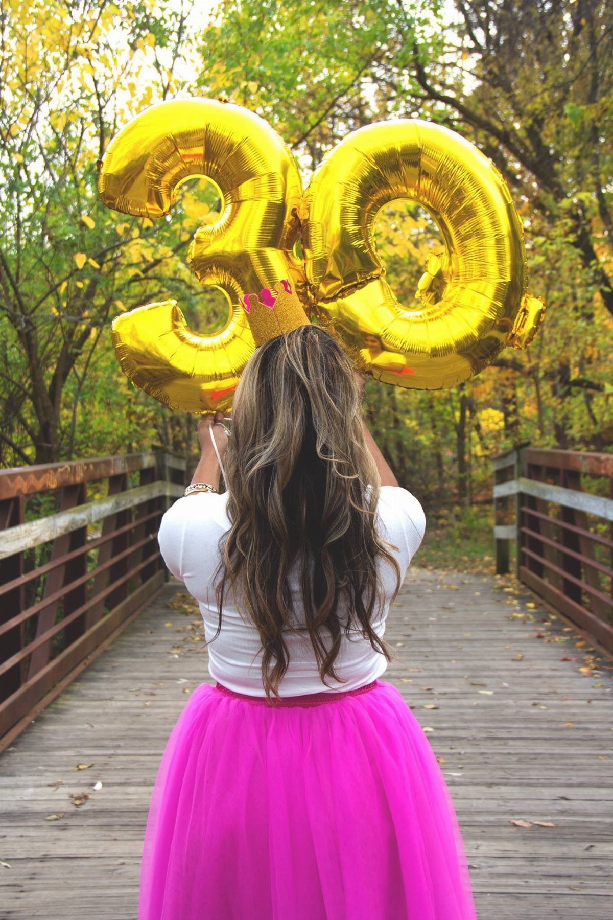 Woman in a pink tulle skirt holding large gold number 30 balloons on a wooden bridge.
