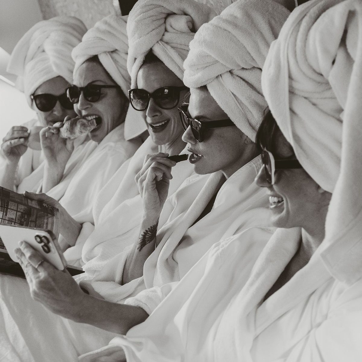 Five women in white robes and hair towels eating pastries and looking at a phone.
