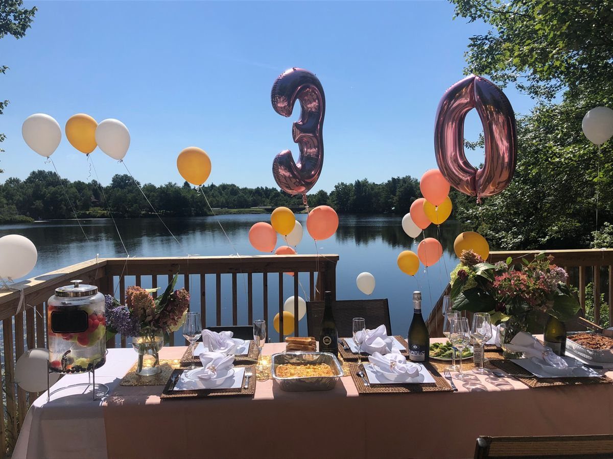 A decorated outdoor dining table with rose gold 30 balloons overlooking a peaceful lake.