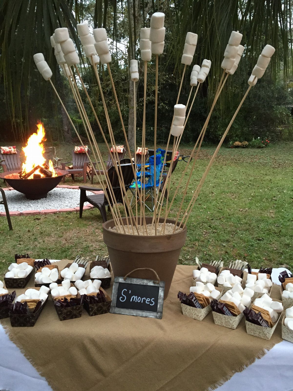 S'mores dessert spread with marshmallows on skewers and chocolate treats in baskets outdoors.