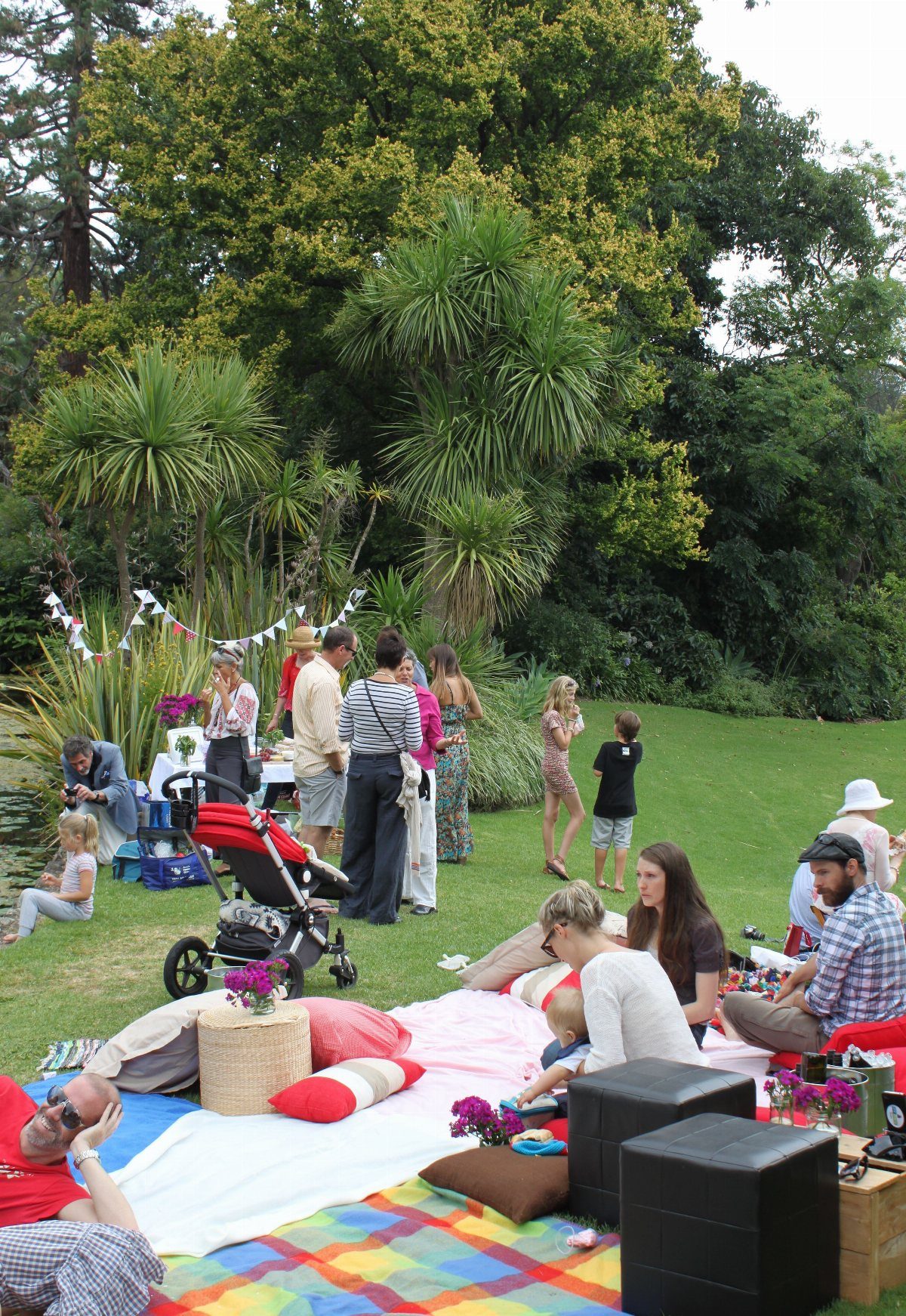 People relaxing on blankets and cushions during an outdoor gathering in a lush green park.