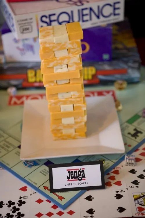 A tower of cheese cubes stacked like Jenga on a white plate over game pieces.