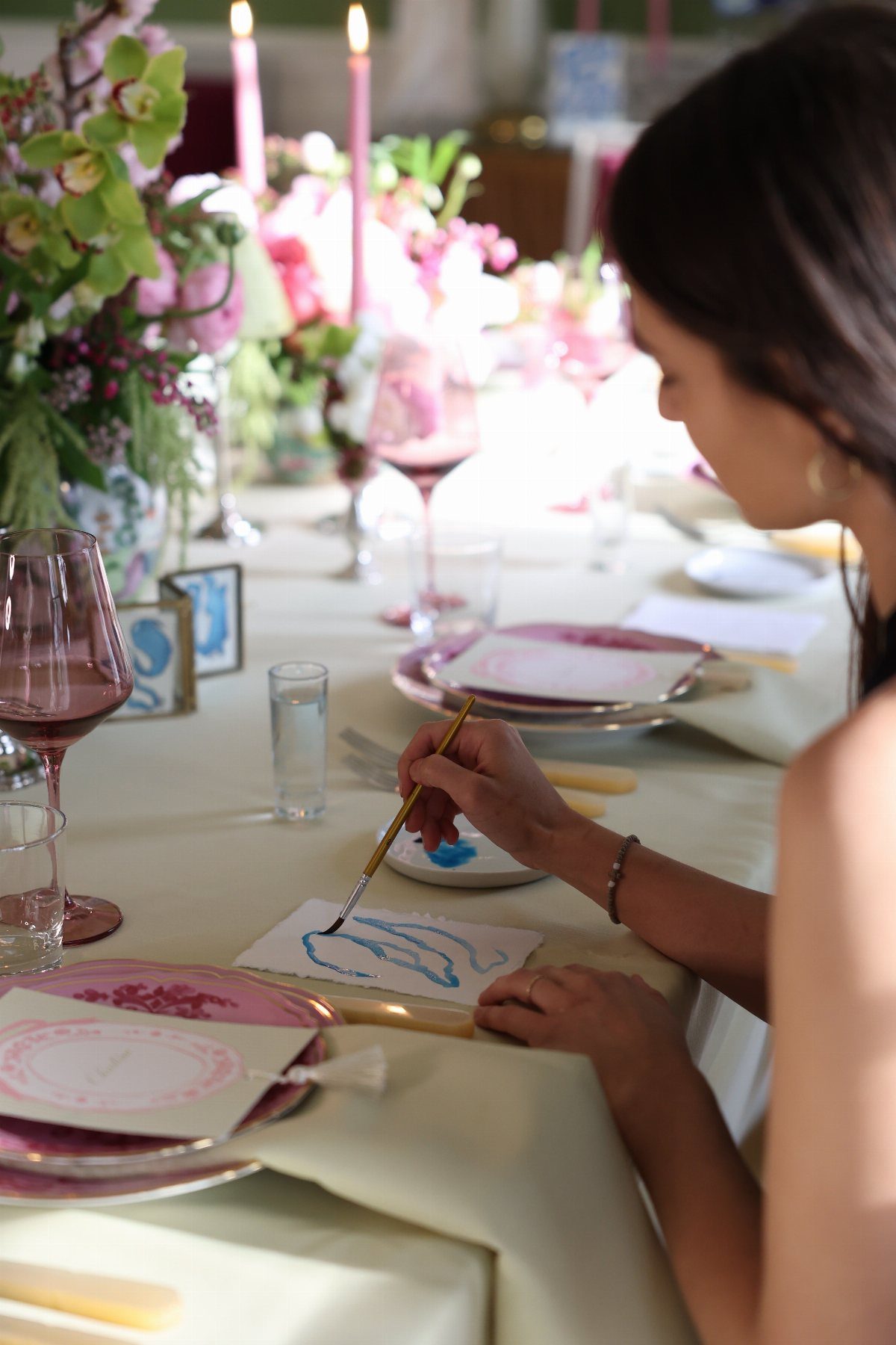 A woman painting on small paper at an elegantly set dining table with flowers.