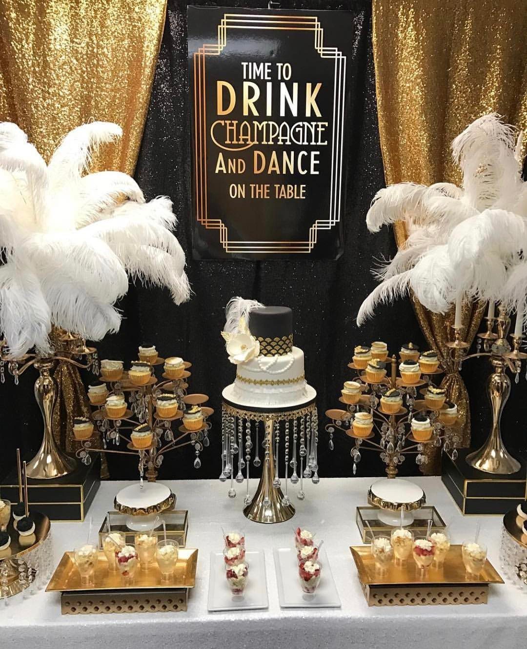 A glamorous dessert table with a white cake, gold cupcake stands, and white feather plumes.
