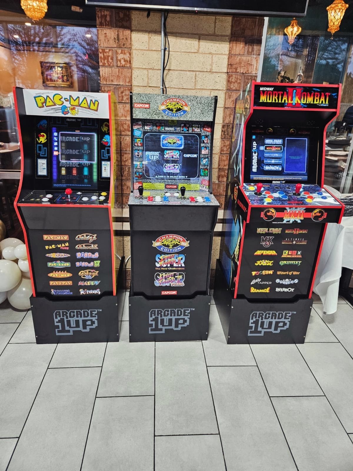 Three colorful arcade game cabinets including Pac-Man and Mortal Kombat standing on a tiled floor.