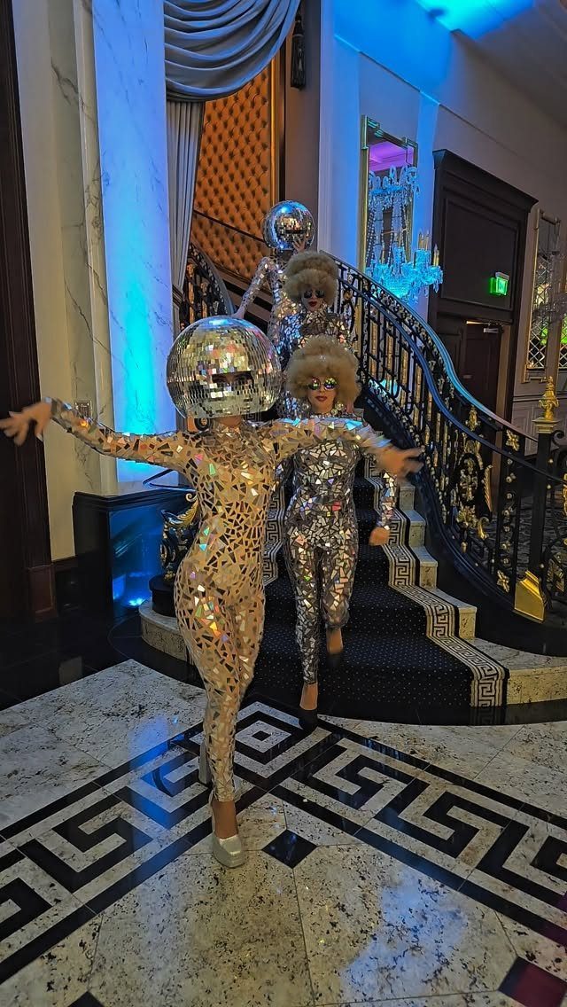Performers in silver sequined bodysuits and disco ball helmets posing on a grand staircase.