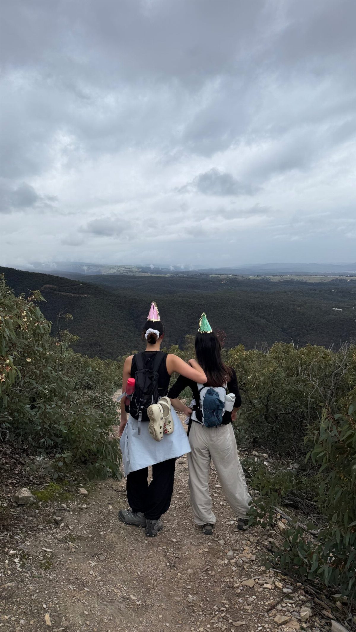 Two women wearing party hats and backpacks stand on a mountain trail overlooking a valley.