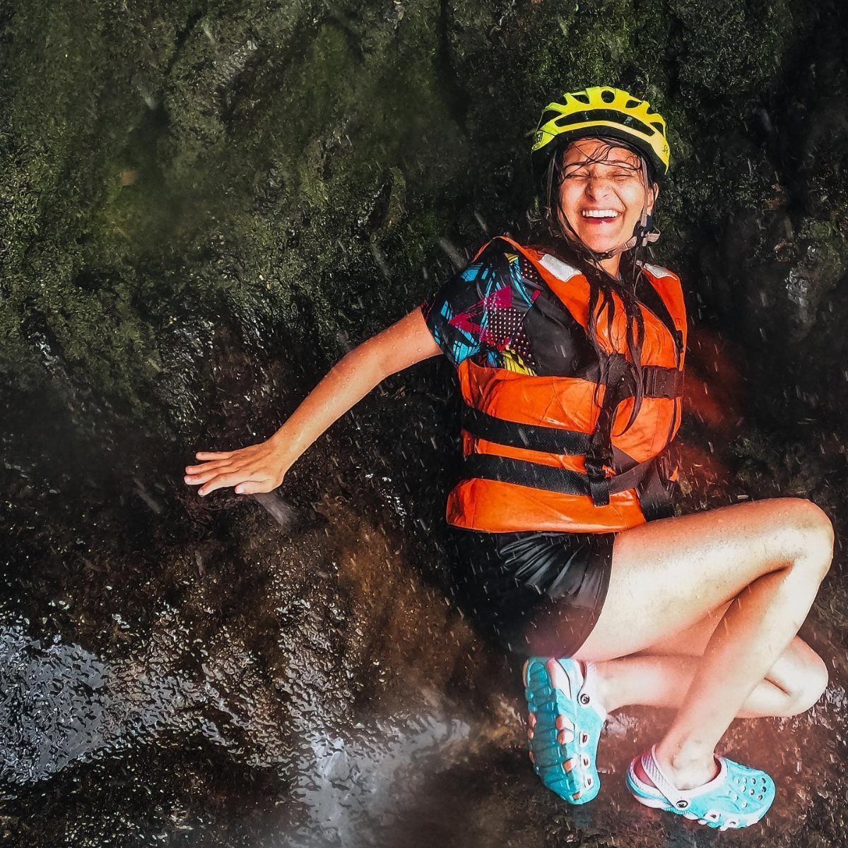 A smiling woman in a life jacket and helmet sitting on wet rocks near water.
