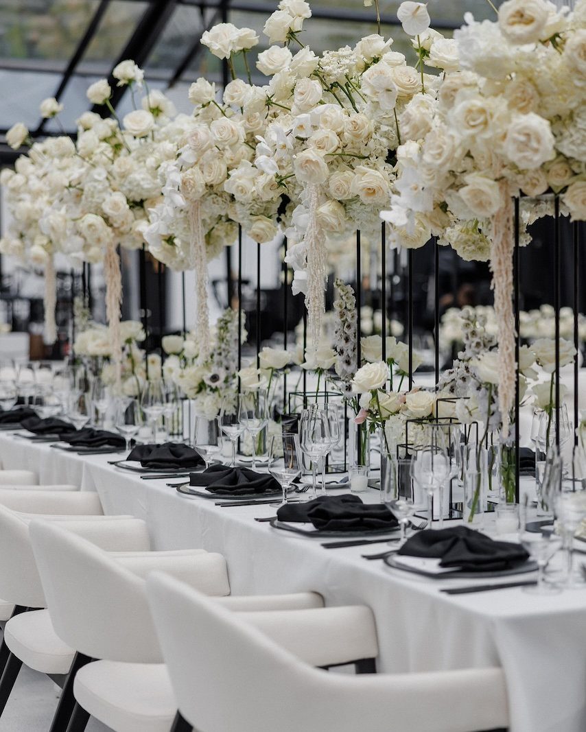 Elegant dining table set with white linens, black napkins, and tall white floral arrangements.