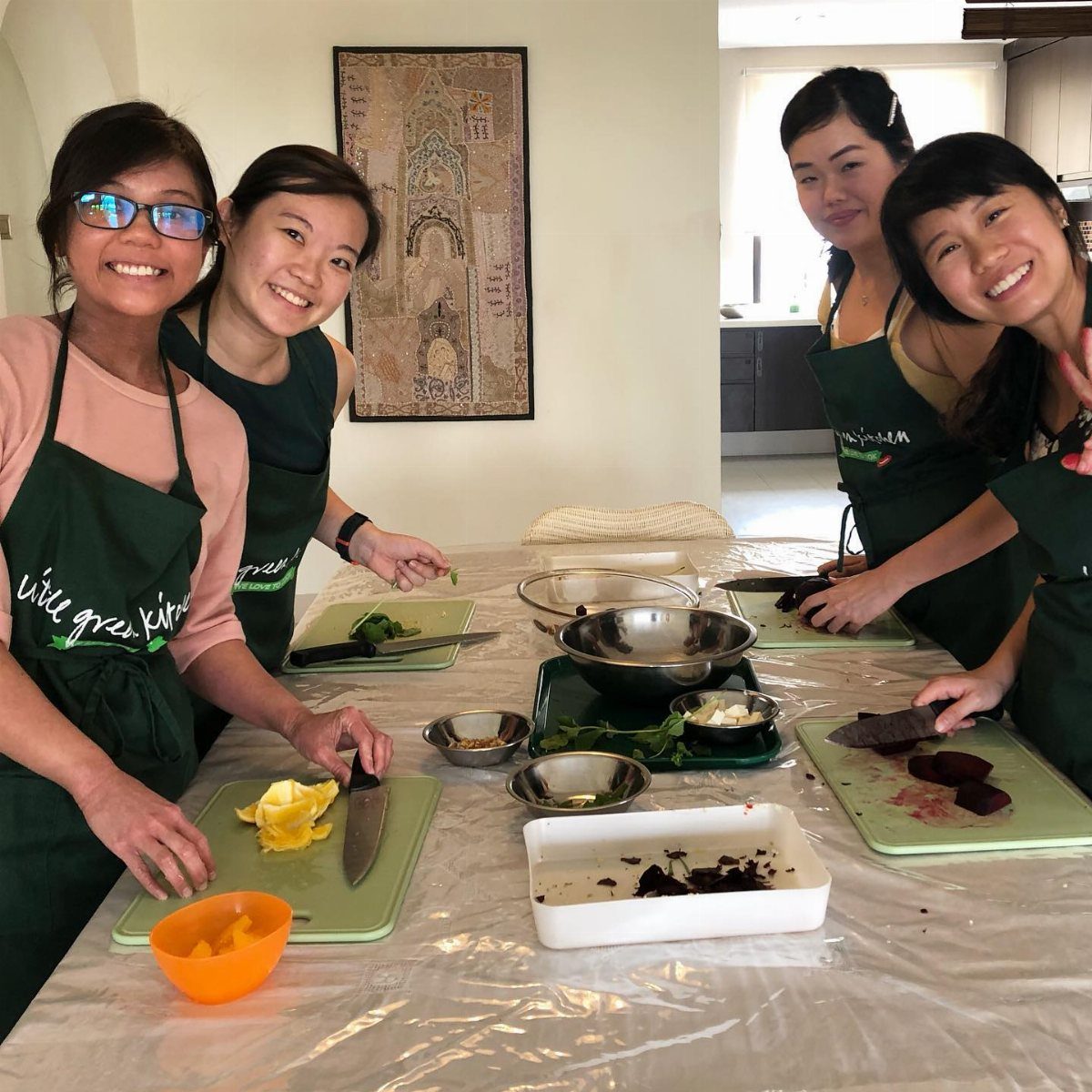Four women in green aprons smiling while preparing vegetables at a kitchen table.