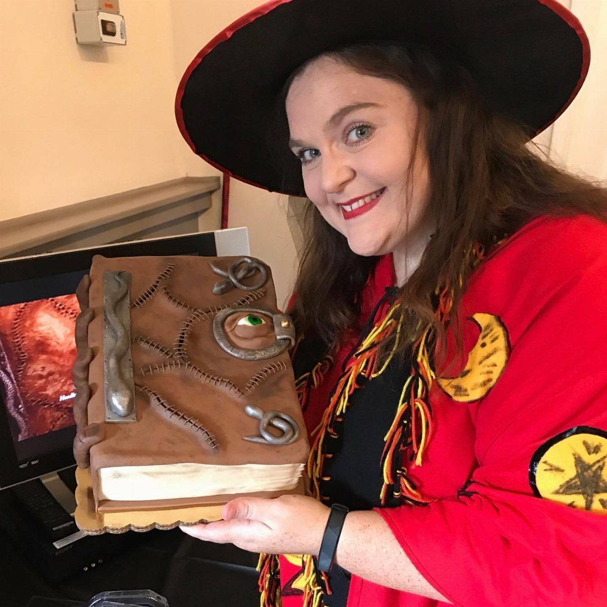 A woman in a witch costume smiling while holding a cake shaped like an old book.