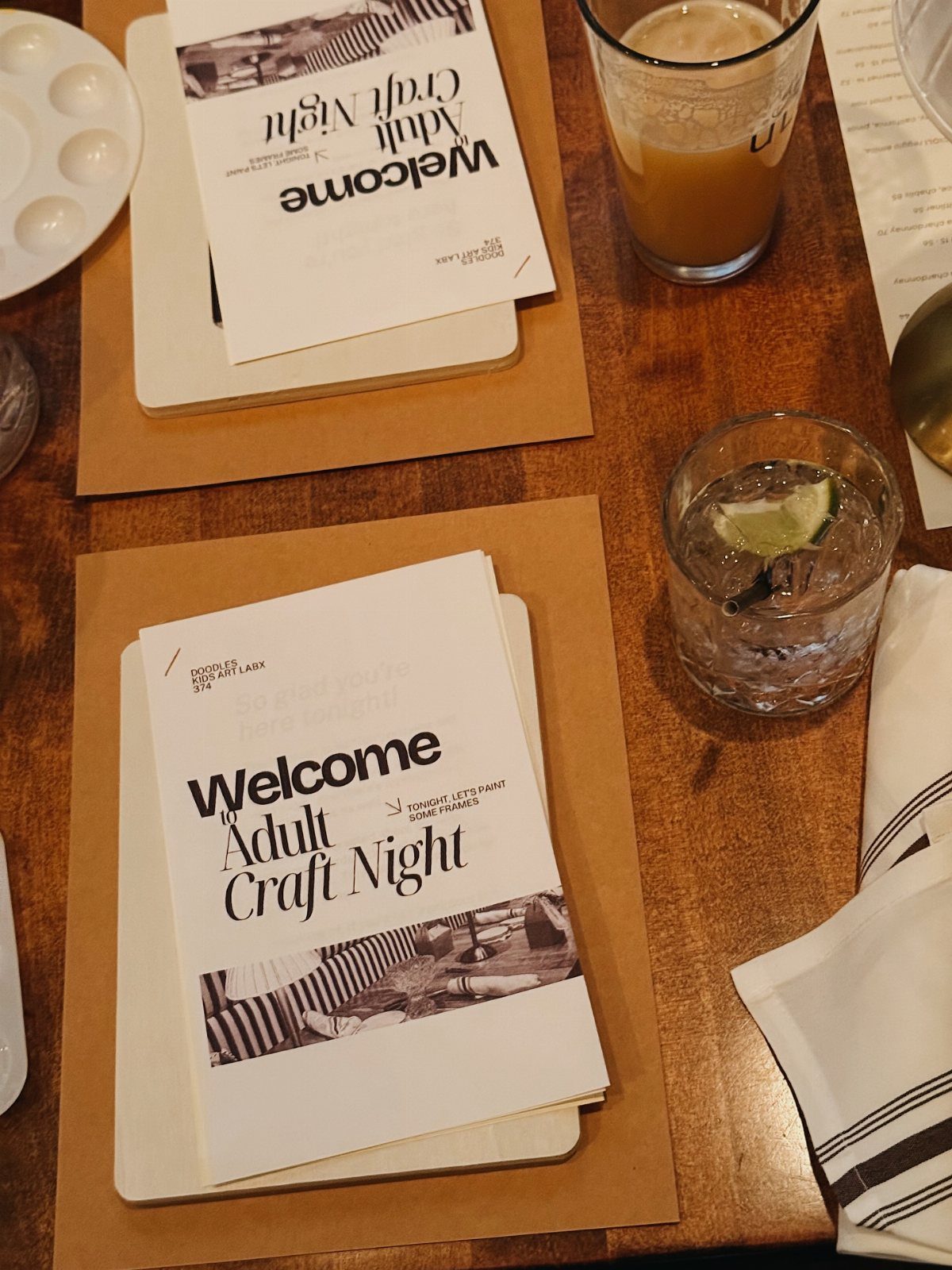 Two welcome booklets for an adult craft night sitting on a wooden table with drinks.