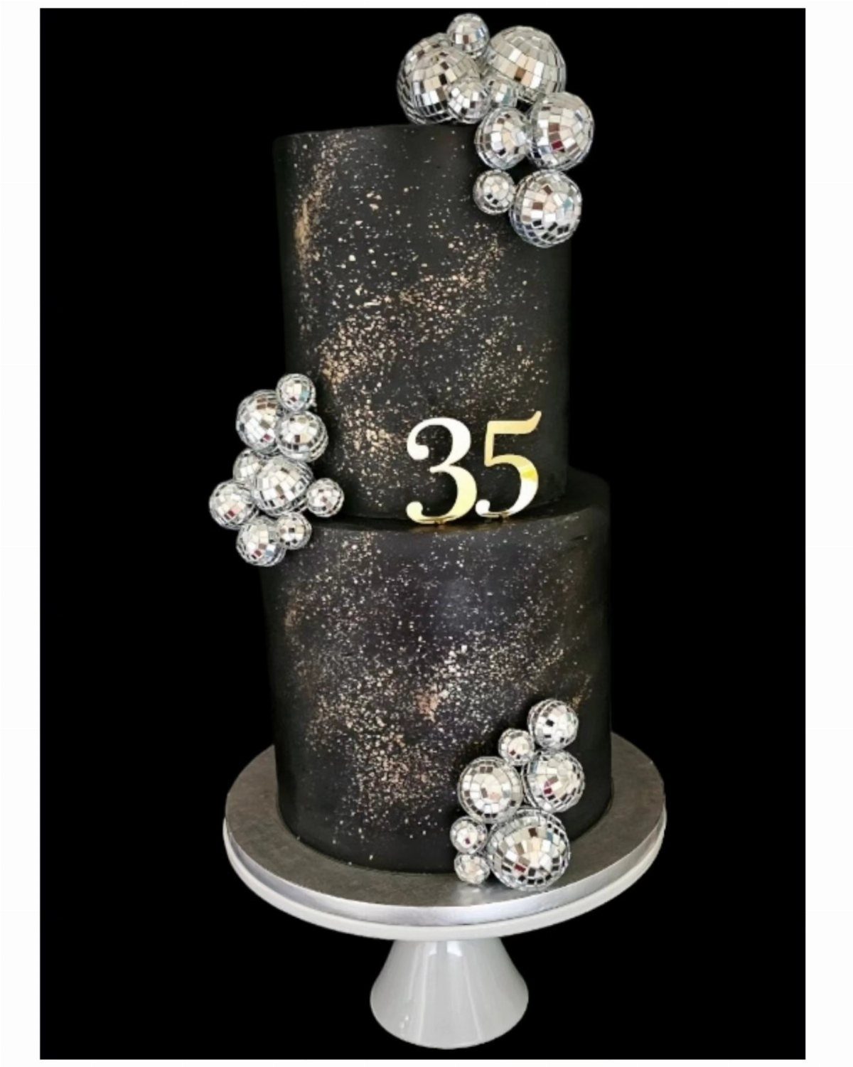 A two-tier black cake with gold speckles, silver disco ball decorations, and a gold number 35.