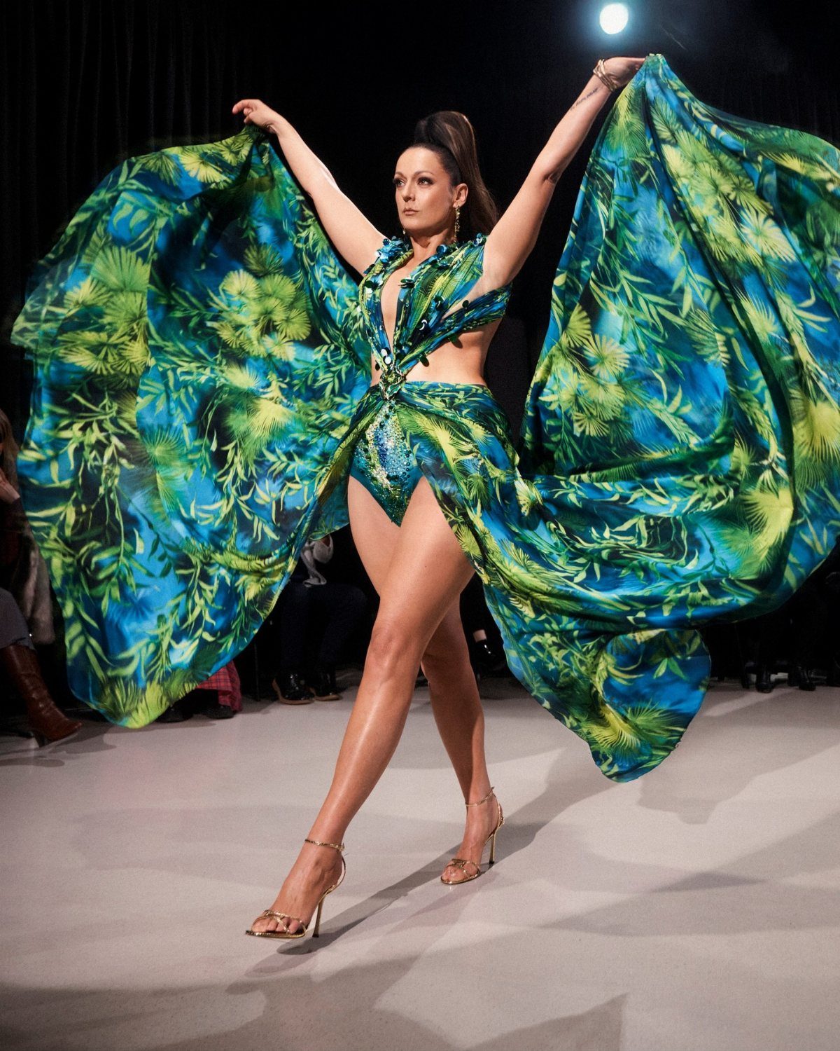A model in a tropical print gown walks a runway with arms raised.