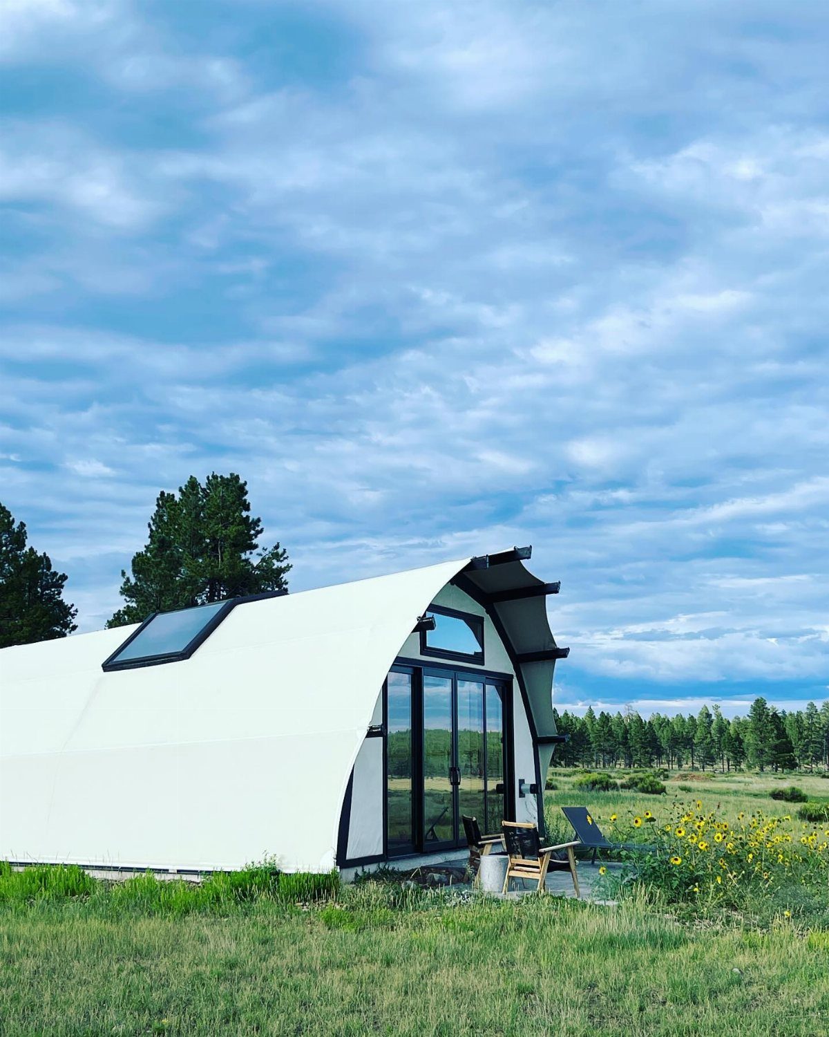 A modern white curved glamping pod situated in a lush green meadow under a cloudy sky.