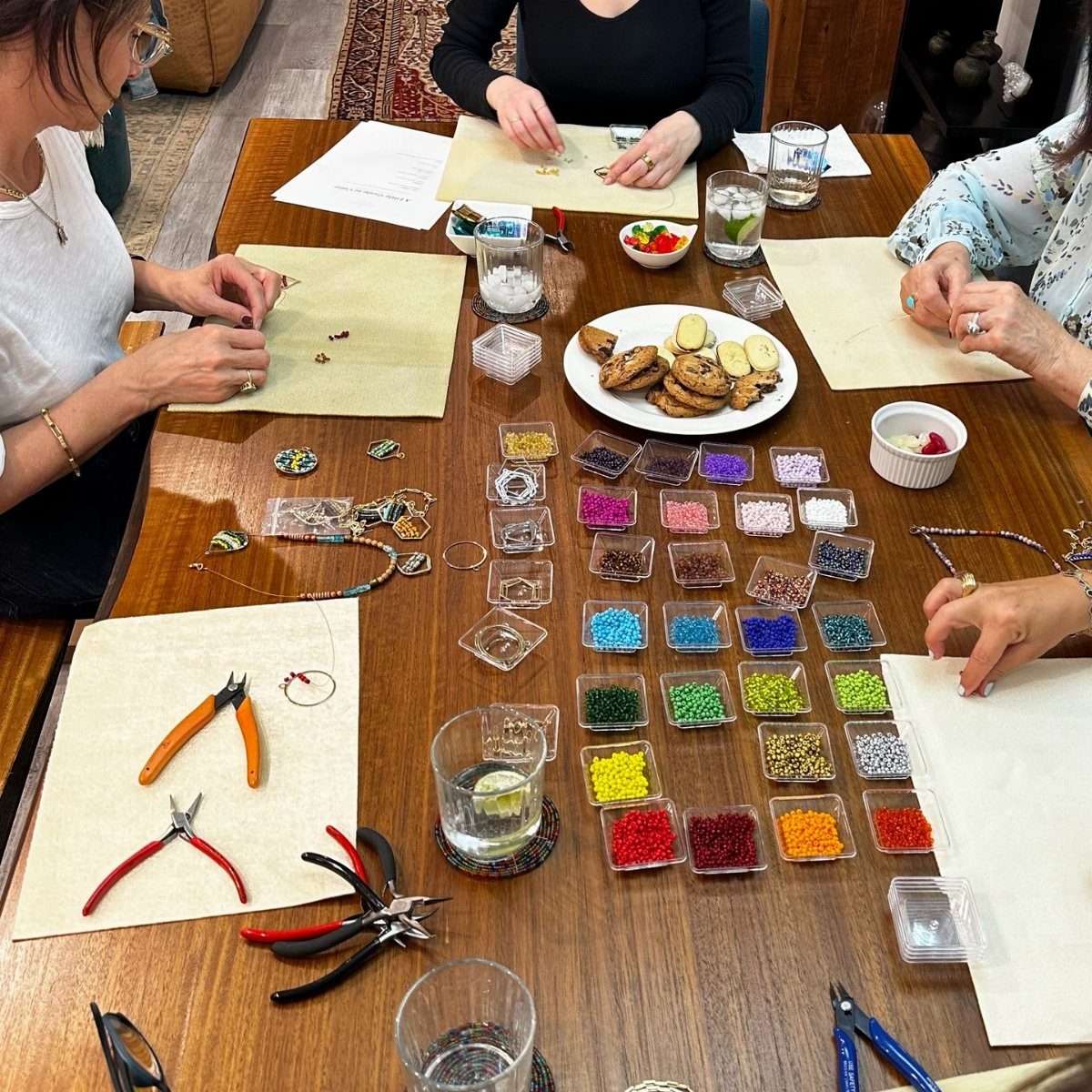 Women sitting at a wooden table assembling jewelry with colorful beads and pliers.