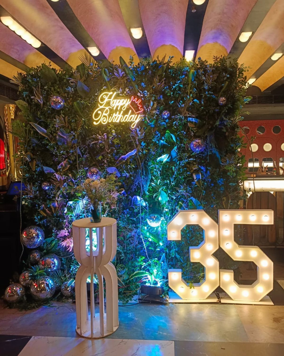 A green foliage wall with disco balls, a neon birthday sign, and large lighted number 35.