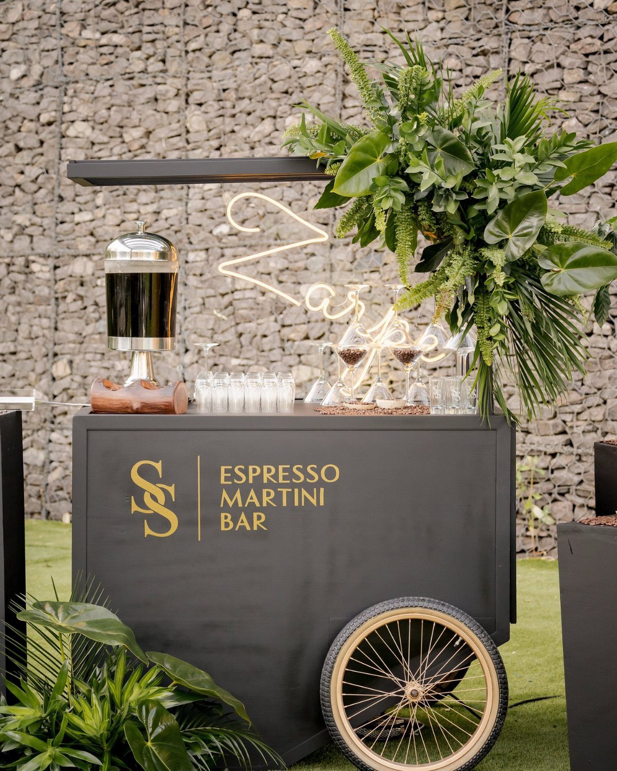 A black mobile espresso martini bar cart decorated with tropical green leaves and gold lettering.