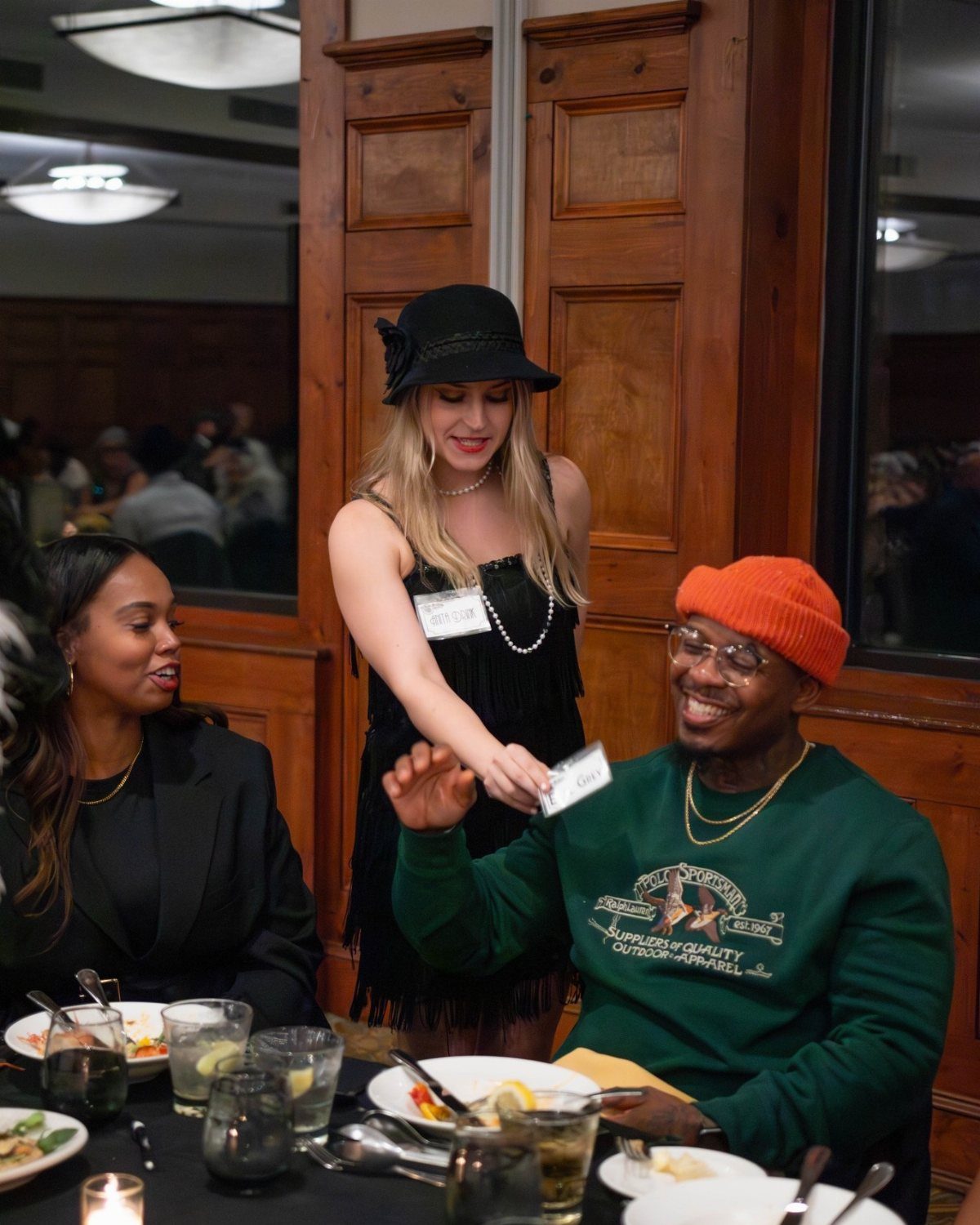 A woman in a black hat hands a small card to a man at a dinner table.