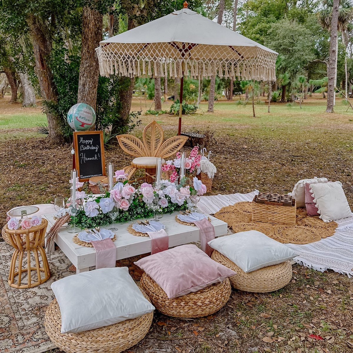 A decorated low picnic table with pink flowers and pillows set outdoors under a fringed umbrella.