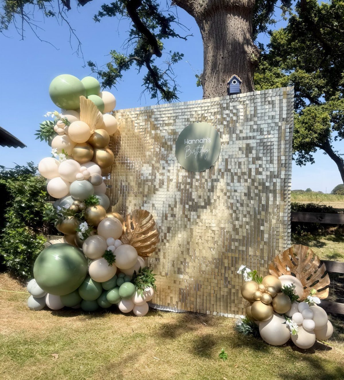 Gold sequin backdrop decorated with sage green and cream balloon garlands and palm leaves outdoors.