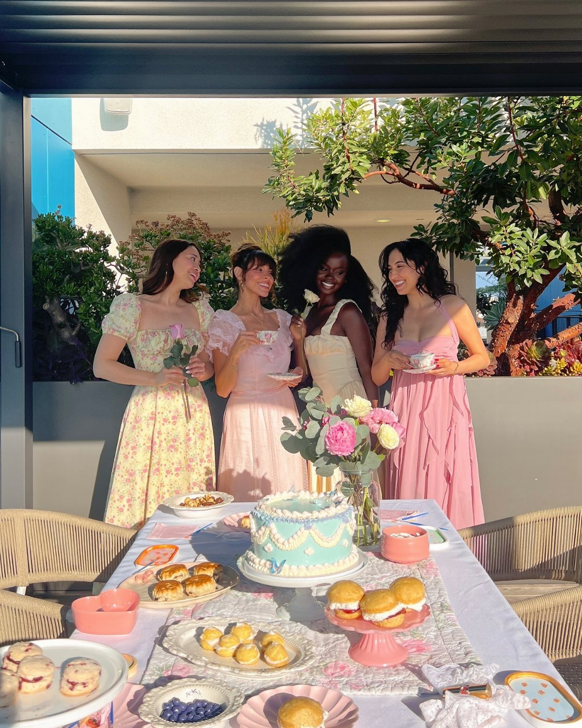 Four women in pastel dresses gather around a table filled with tea, pastries, and a blue cake.
