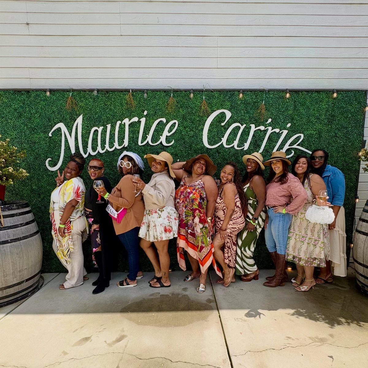 Ten women posing in front of a green hedge backdrop with white cursive lettering.