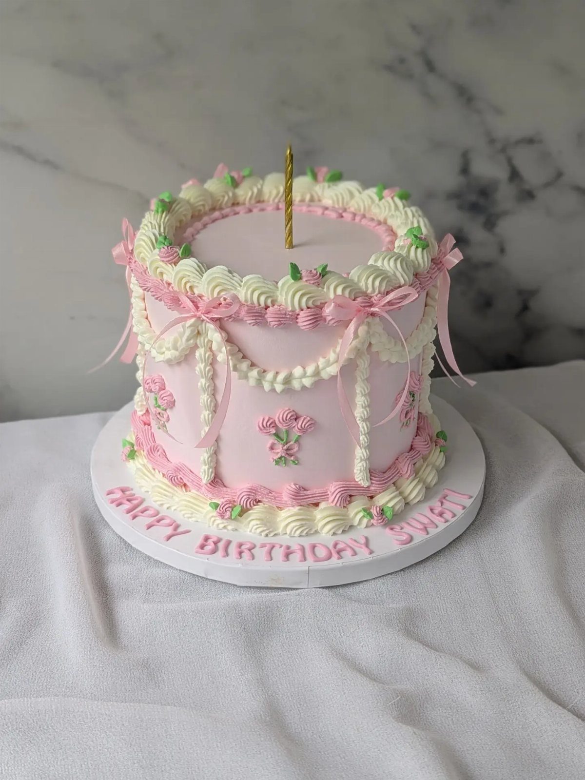 Pink frosted cake with white buttercream swirls, small floral decorations, and pink bows on a white board.