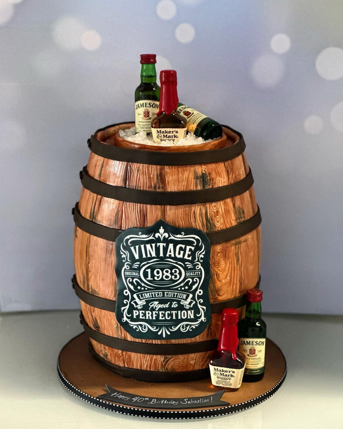 A whiskey barrel shaped cake decorated with miniature liquor bottles and a vintage label.