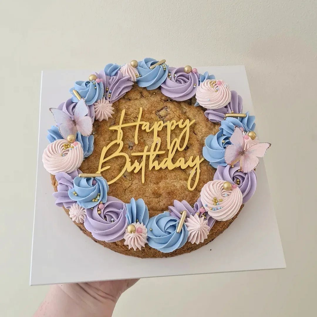 Large chocolate chip cookie cake decorated with pastel frosting swirls, gold lettering, and butterflies.