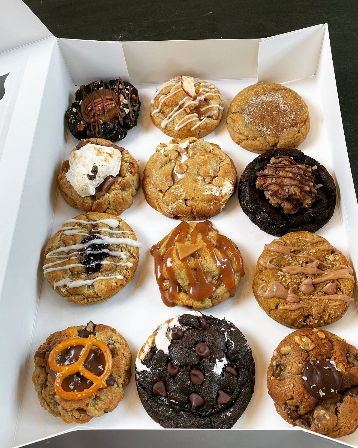Assorted gourmet cookies with various toppings arranged inside a white cardboard box.