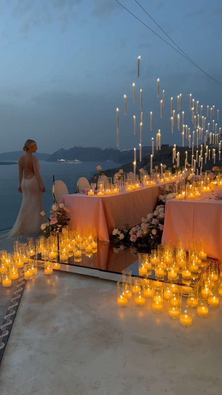A long pink dining table decorated with many glowing candles overlooking the ocean at dusk.