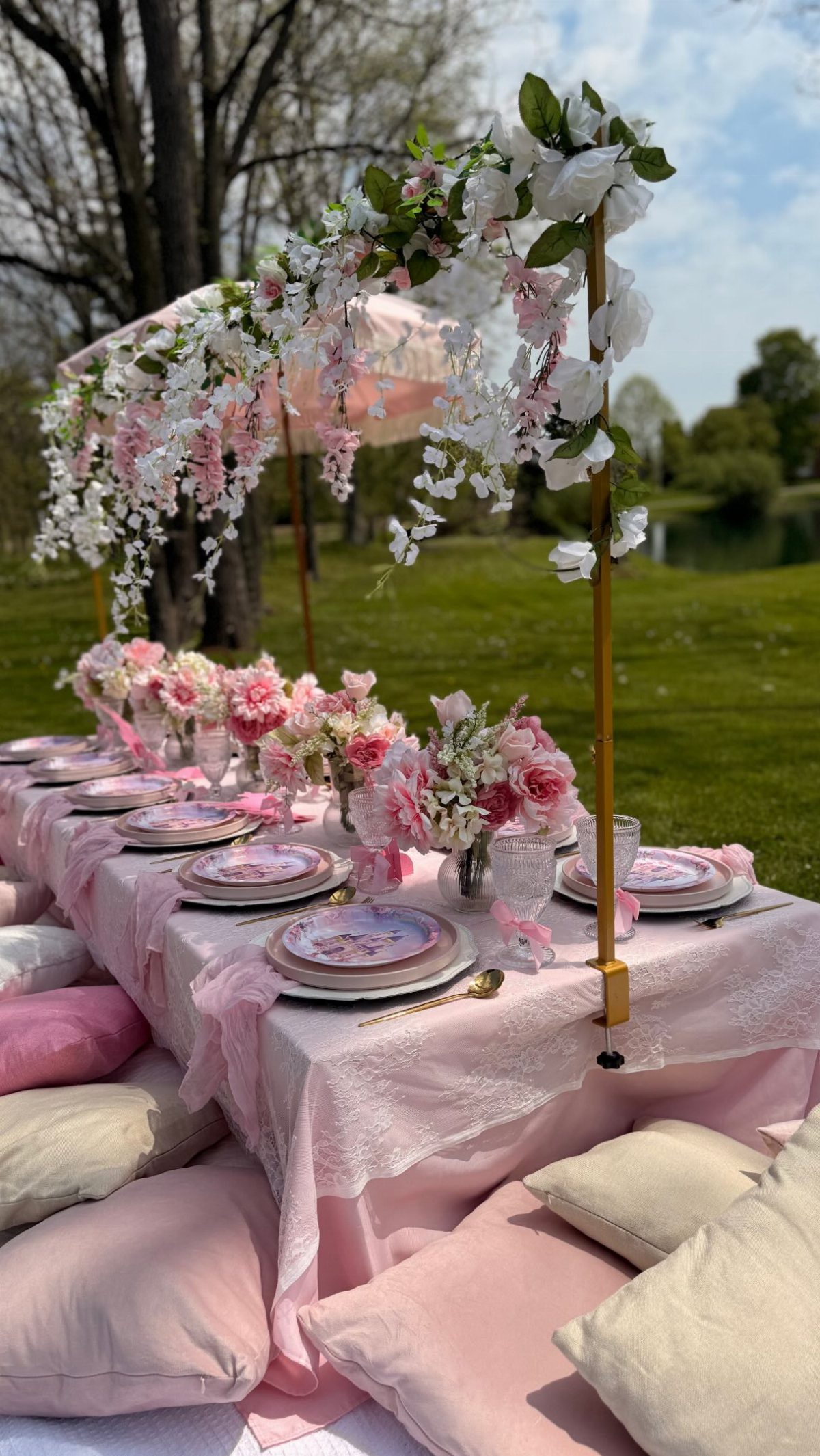 A low picnic table decorated with pink flowers, lace, and cushions on a green lawn.