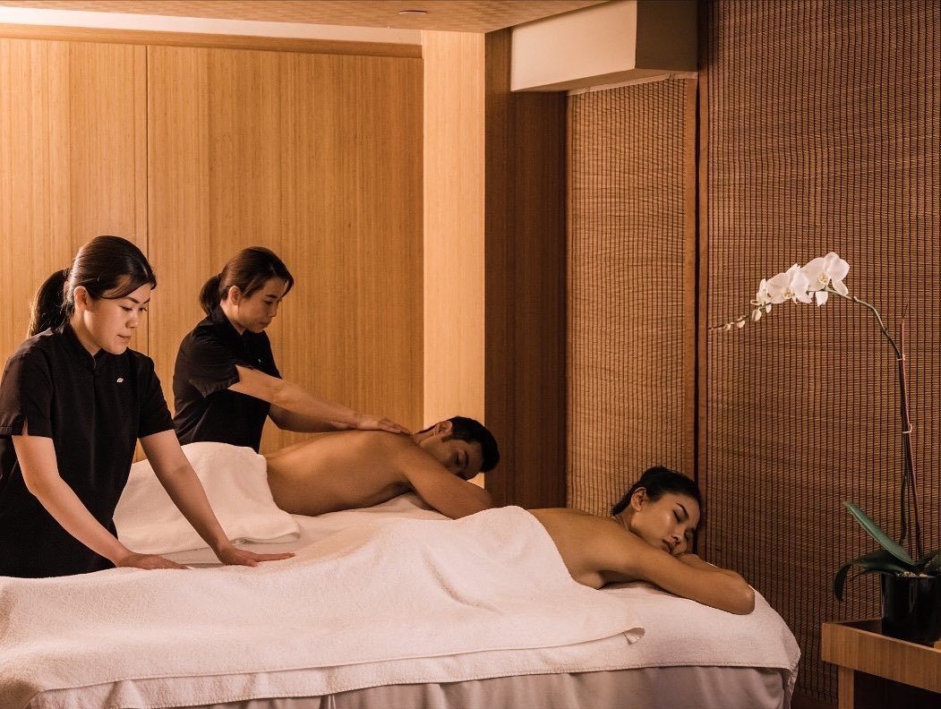 Two massage therapists performing a treatment on two men in a warm spa setting.