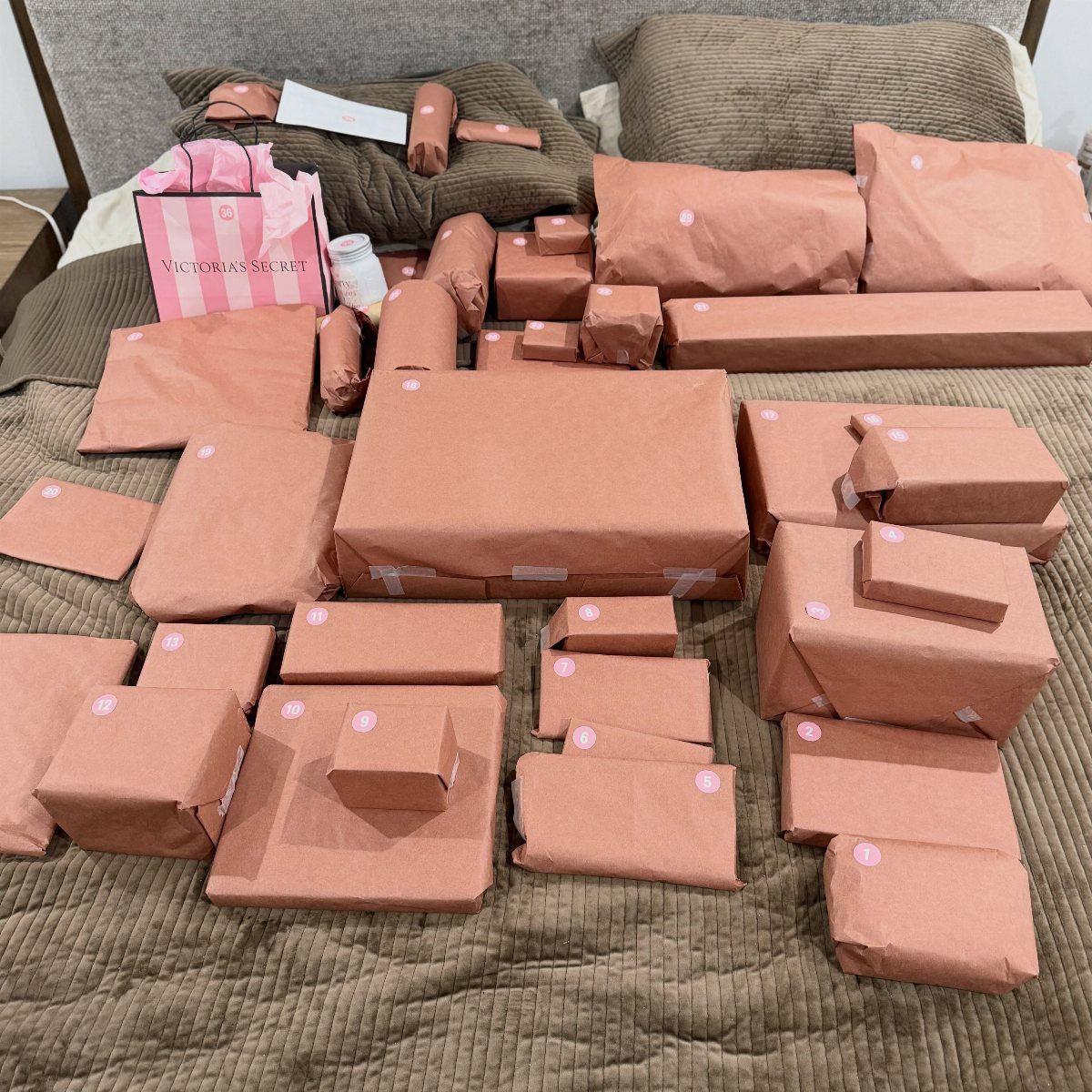 Many brown paper wrapped gifts spread across a brown quilted bed with a pink gift bag.