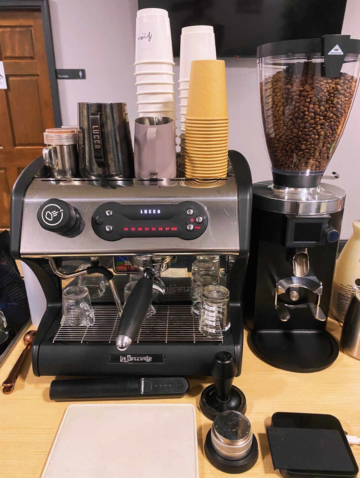 A silver and black espresso machine with coffee beans in a grinder and stacks of cups.