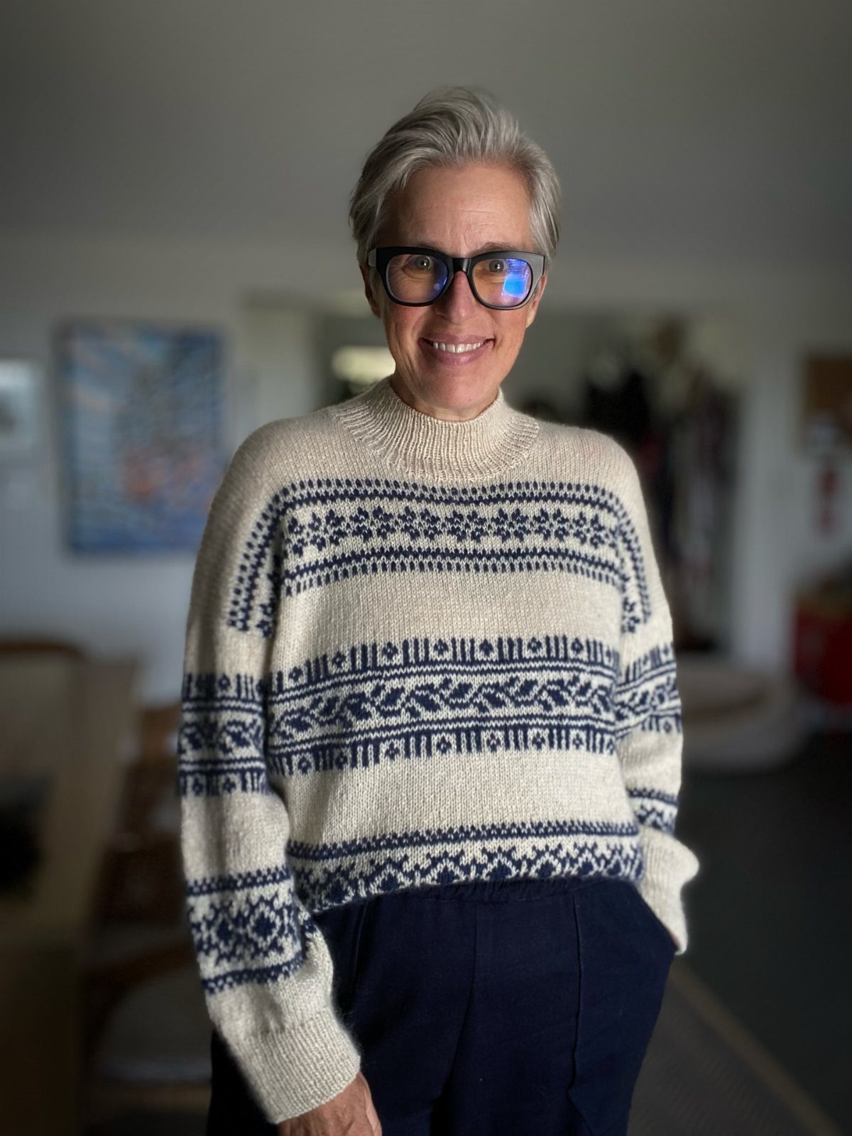 A smiling woman with grey hair wearing a cream and blue patterned knit sweater.