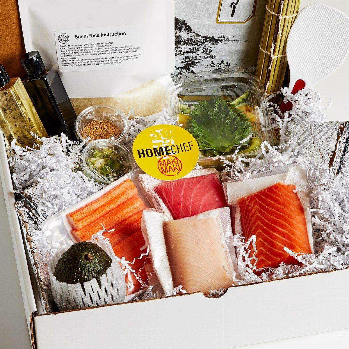 A white gift box filled with fresh sushi ingredients including salmon, tuna, avocado, and rice instructions.