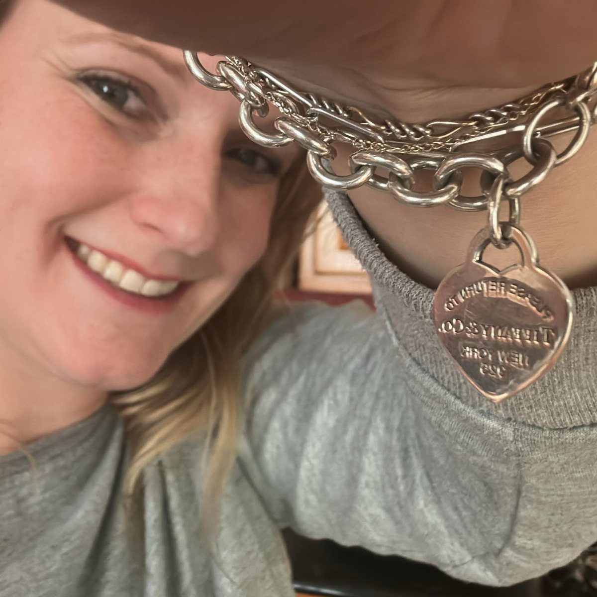 A smiling woman holding up a silver heart charm bracelet towards the camera.