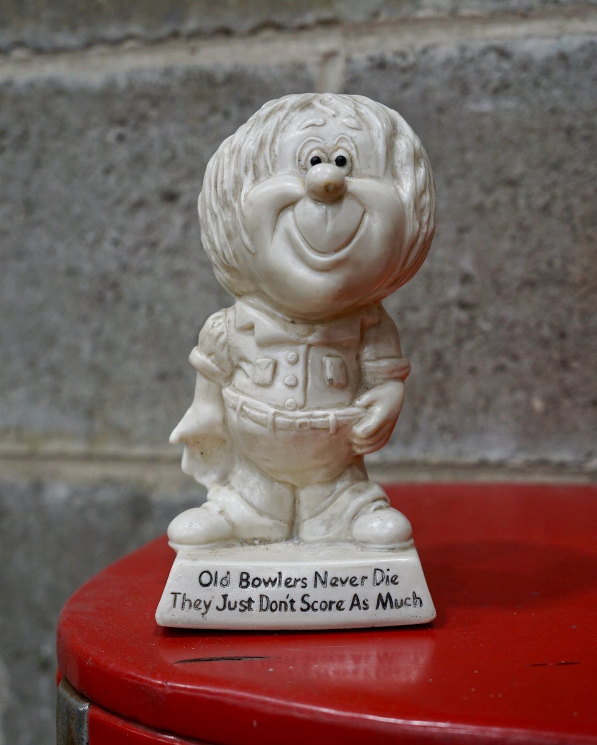 A white ceramic bowling-themed figurine sitting on a red surface against a stone wall.