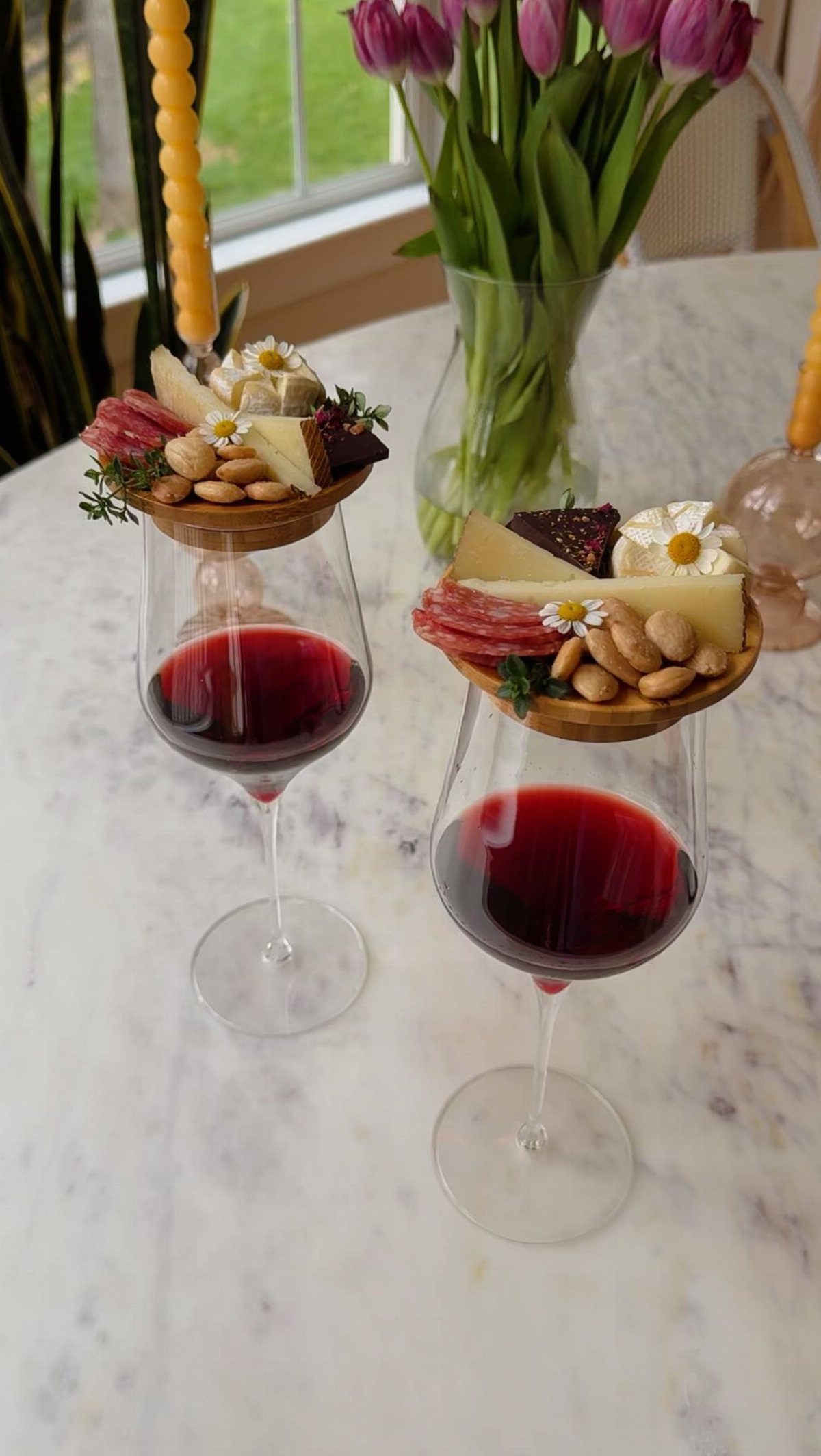 Two wine glasses filled with red wine topped with small charcuterie platters on a marble table.