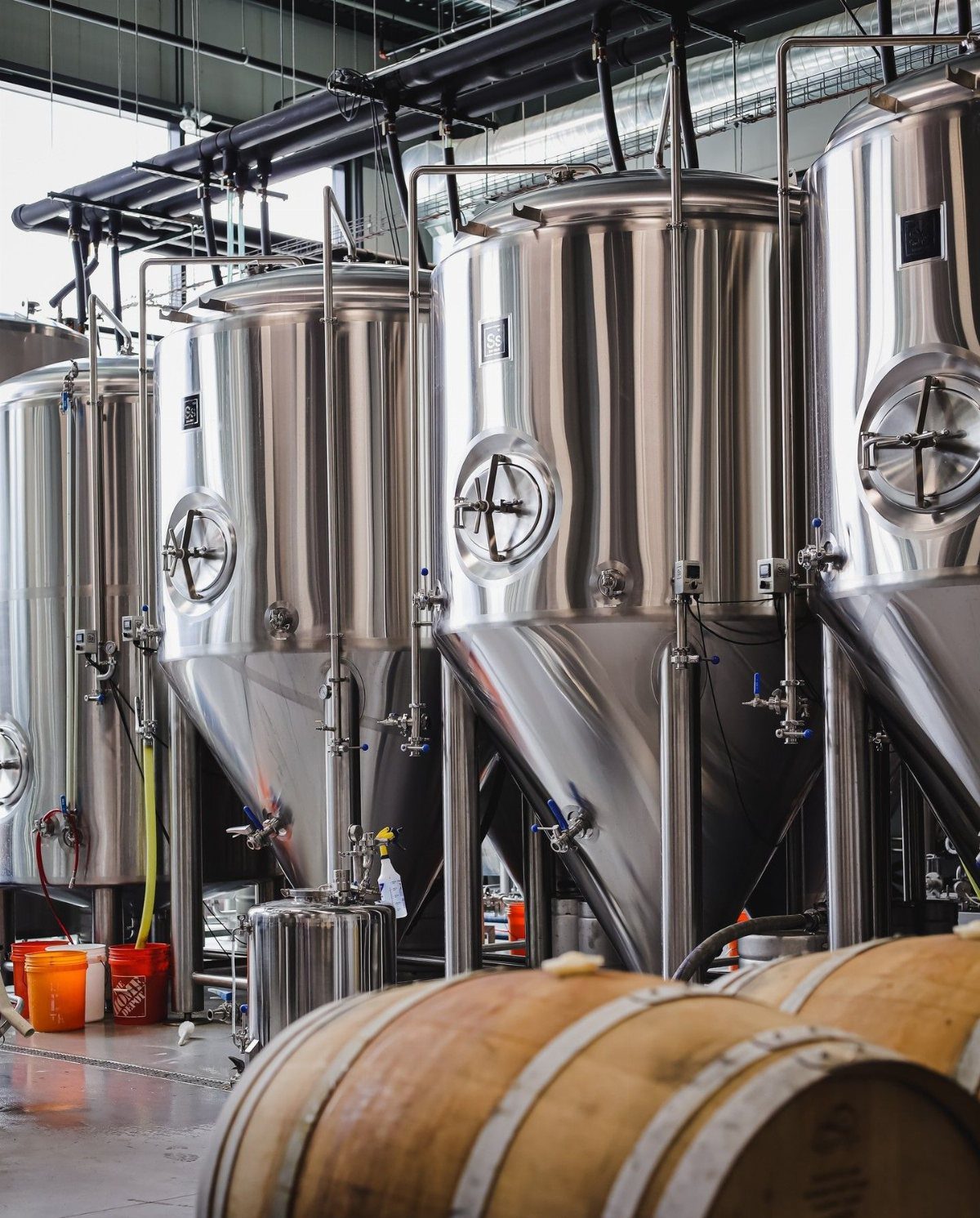 Large stainless steel brewery tanks and wooden barrels in a production facility.