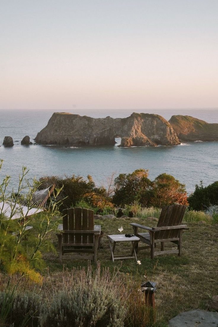 Two wooden chairs and a small table overlooking a scenic ocean view with large rocks.