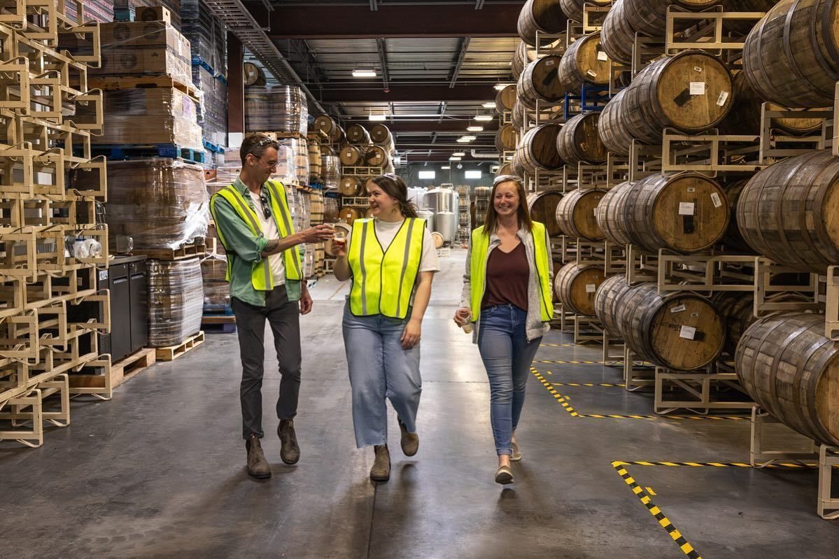 Three people in high-visibility vests walking through a warehouse filled with wooden barrels.