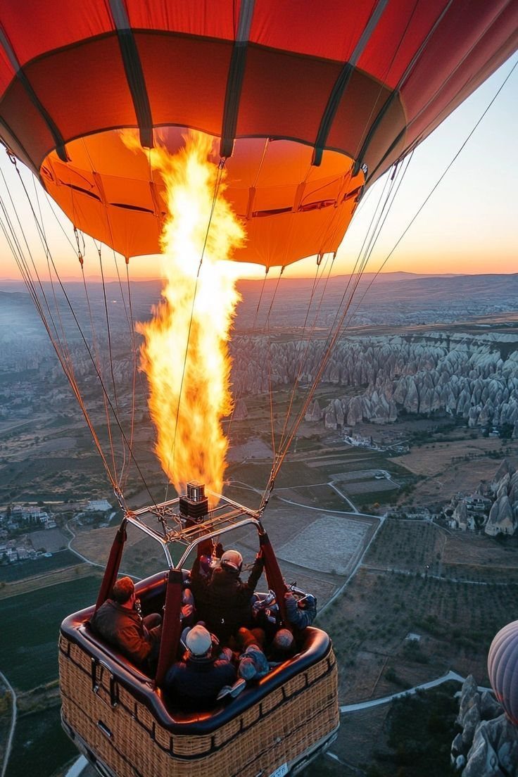 A red hot air balloon with a large flame soaring over a scenic landscape at sunset.
