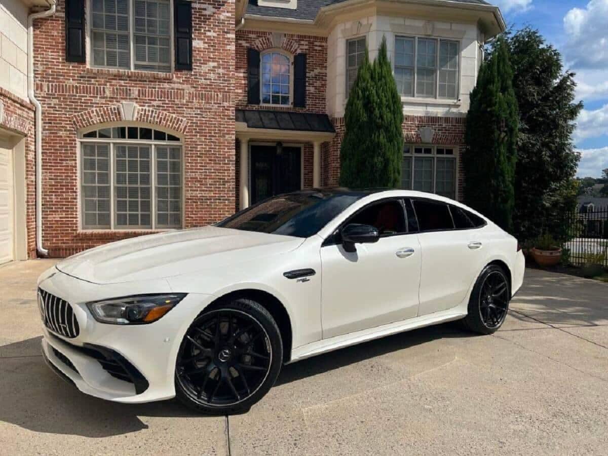 A white luxury Mercedes sedan parked on a concrete driveway in front of a brick house.